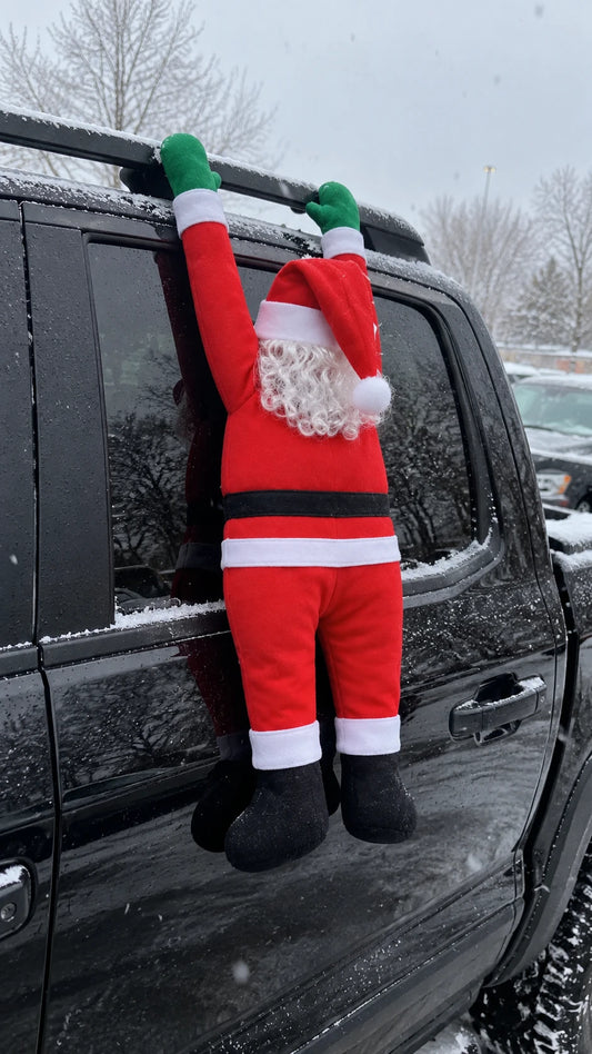 Hanging Santa draped over the roof rack of a black pickup truck in a snowy car park, arms up and boots dangling beside the window